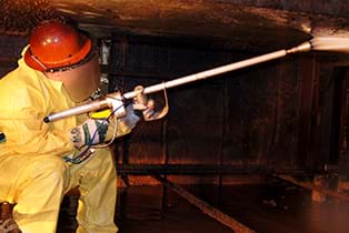 A worker covered in safety gear is using a high-pressured water jet to remove hardened materials from a rail car