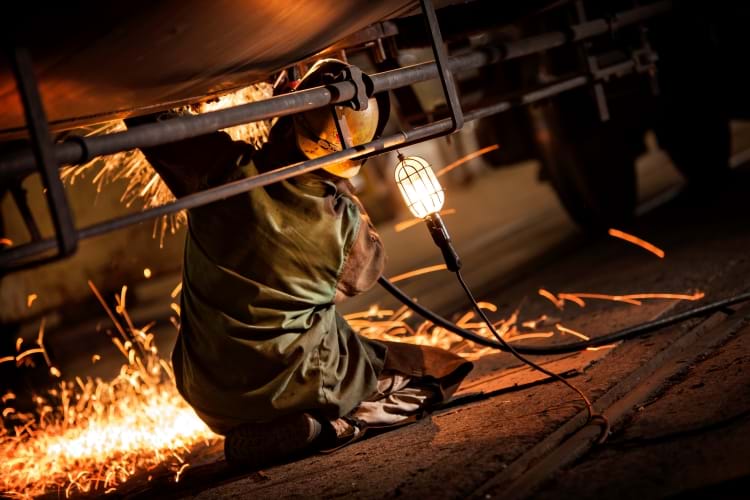 A Welder on the floor of a railcar repair shop welding the underside of a tank car