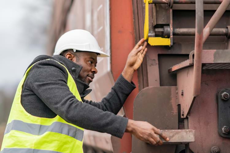 A mechanic wearing safety equipment (helmet and jacket) inspecting components of a freight train
