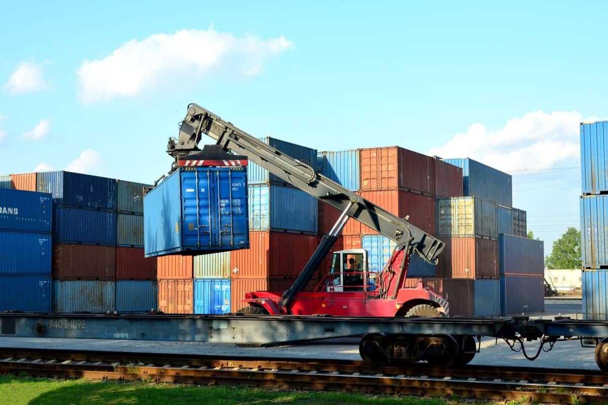 Container handler loading an intermodal container onto a rail car beside container stacks