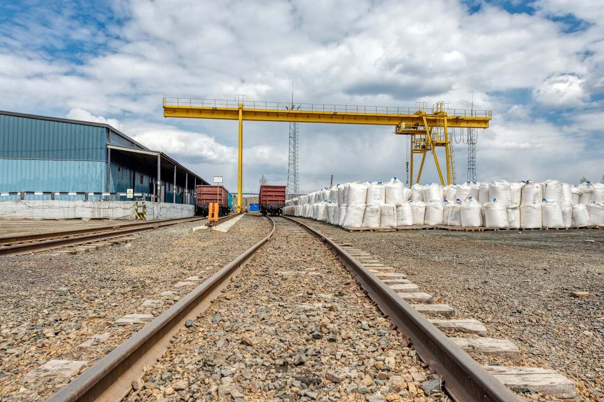 Transload yard: railcars on a siding beside a warehouse, with a gantry crane over bagged freight