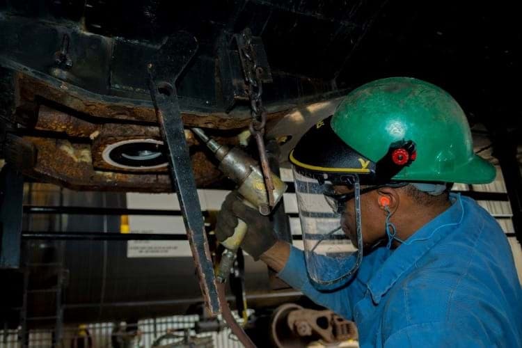 A shop worker in a hard hat and face shield working on the underside of a freight railcar