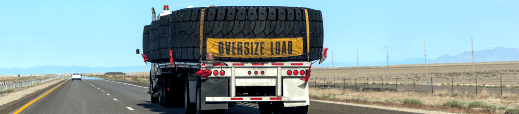 Large tires are being transported on their side on a flat deck trailer. An oversized load banner hangs from the back of the trailer.